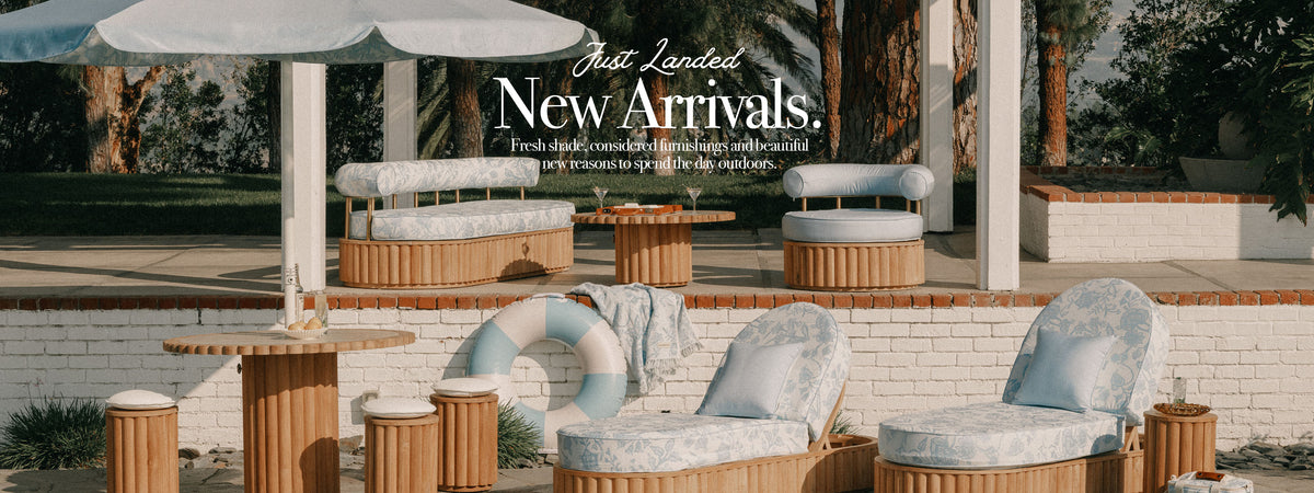 Outdoor patio furniture arrangement with light blue patterned cushions, round wooden tables, and a white brick wall in a landscaped garden setting. Large overlaid text reads 'Just Landed New Arrivals.