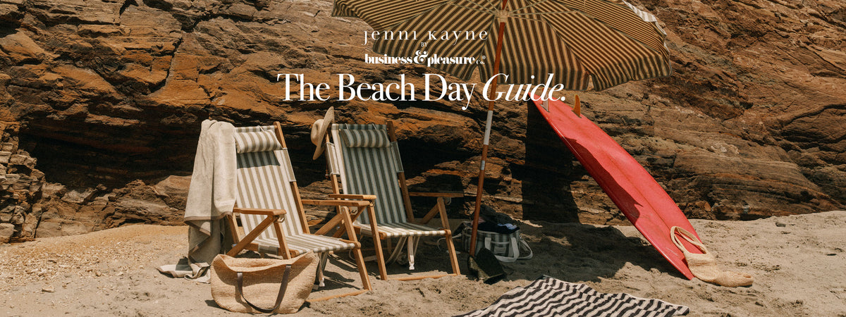 A beach day scene set against a large rock cave, featuring two striped wooden deck chairs, a striped beach umbrella, a red surfboard leaning against the wall, and a black-and-white striped towel on the sand. Large overlaid text reads 'JENNI KAYNE' and 'The Beach Day Guide.