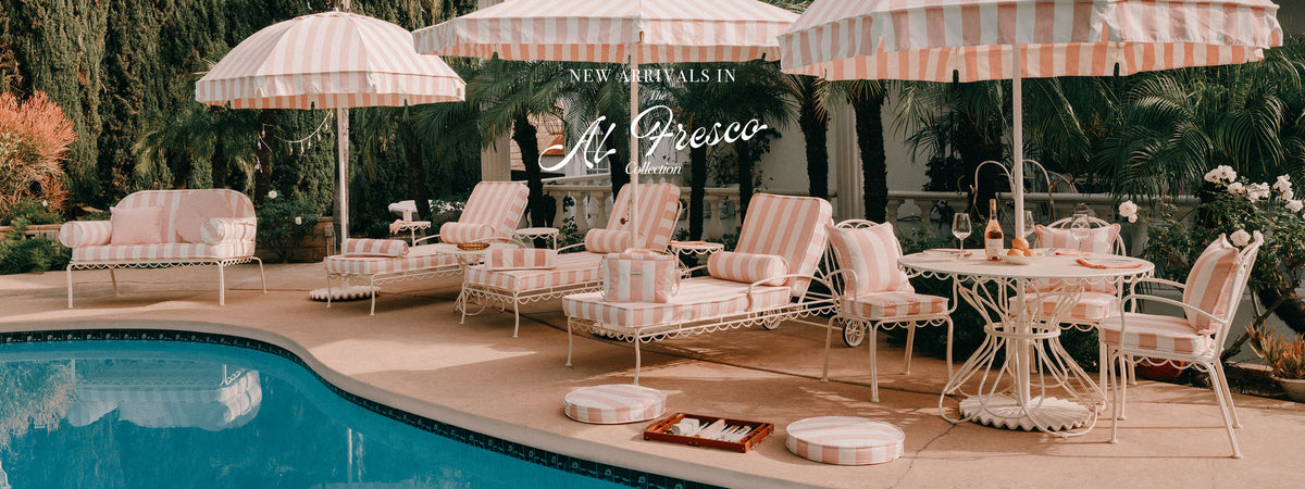 A luxurious outdoor poolside lounge set featuring white wicker chairs and striped pink-and-white umbrellas on a beige patio, with a swimming pool in the foreground and a tropical garden background. A central text overlay reads 'NEW ARRIVALS in Al Fresco'.