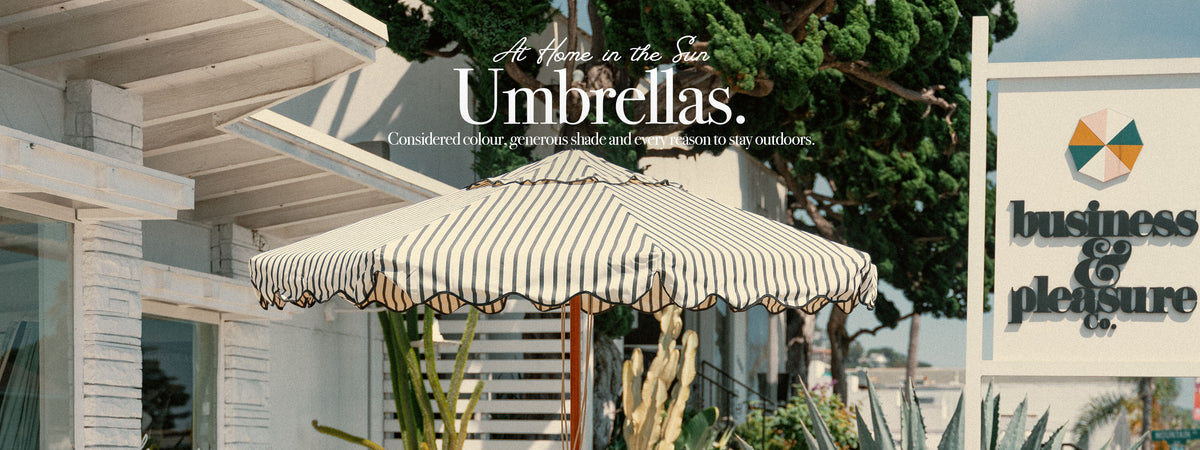A striped patio umbrella in front of a white building with the text 'At Home in the Sun Umbrellas' overlaid, alongside a Business & Pleasure sign on the right.