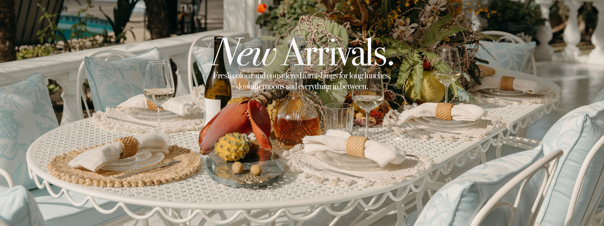 A styled outdoor dining table with white lace tablecloth, white plates, folded napkins, wine glasses, and a decorative centerpiece, overlaid with the text 'New Arrivals.' The scene is set in a garden patio environment with light blue chair covers and warm natural lighting.