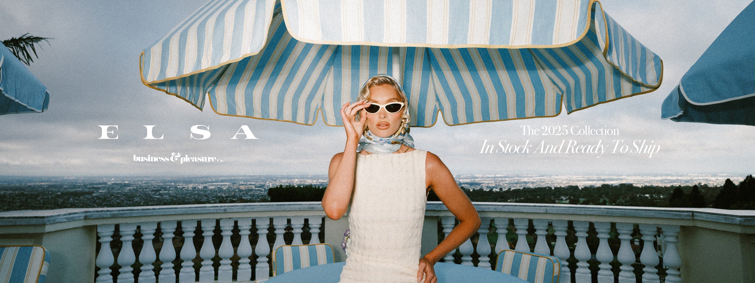 Elsa Hosk in a white dress and sunglasses standing under a large striped umbrella from the 2025 B&P Co. x ELSA Collaboration Collection