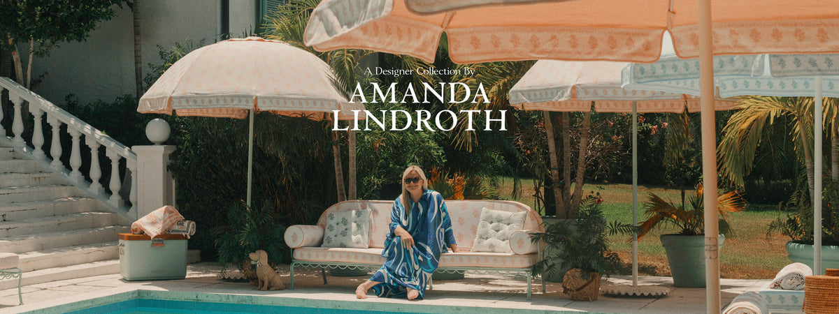 A woman sits by a poolside patio under large cream umbrellas, wearing a blue and white patterned robe. A white sofa, potted plants, and a staircase are visible in the background, with the text 'Amanda Lindroth' overlaid in the center.